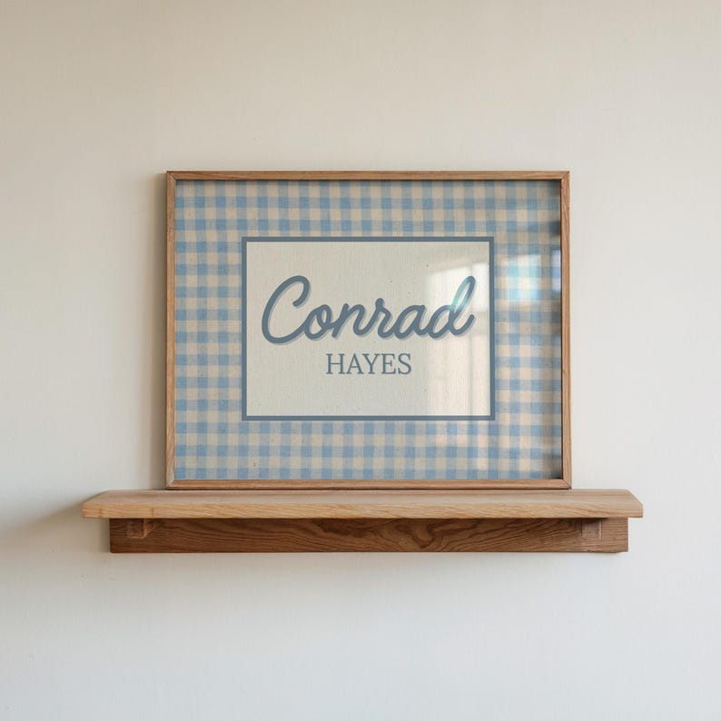 May include: A rectangular wooden frame containing a print with a light blue and white gingham pattern background. Centered on the print is a white rectangular area featuring the name Conrad in a blue script font above the name HAYES in block letters.