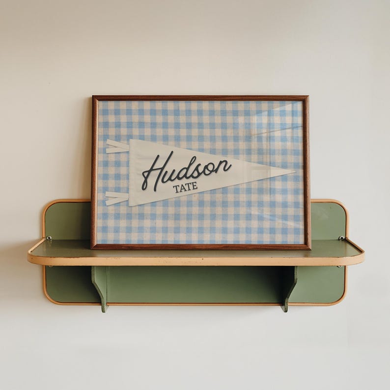 May include: A framed print featuring a white triangular pennant with the text Hudson Tate in black script and block lettering, set against a light blue and white gingham background, displayed within a thin brown wooden frame.