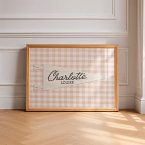 May include: A framed horizontal print featuring a cream-colored triangular pennant with the name Charlotte Louise in dark brown script and block lettering. The pennant is set against a light beige and white gingham checkered background pattern.