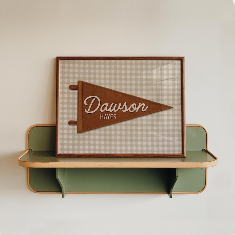May include: A framed brown pennant with the name "Dawson" and the word "Hayes" in a cursive font. The pennant is brown and set against a beige and white checkered background. The frame is brown and the pennant is displayed on a green and beige shelf.