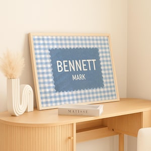 May include: A framed wall art print featuring a light blue and white gingham pattern background with a centered, darker blue, jagged-edged rectangular patch. The text BENNETT MARK is printed in white, sans-serif capital letters on the patch.