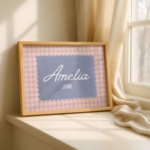 May include: A rectangular wooden frame containing a print with a pink and white gingham background. Centered is a blue fabric-textured patch with pinked edges featuring the white text Amelia and the word JUNE printed in a smaller font below.