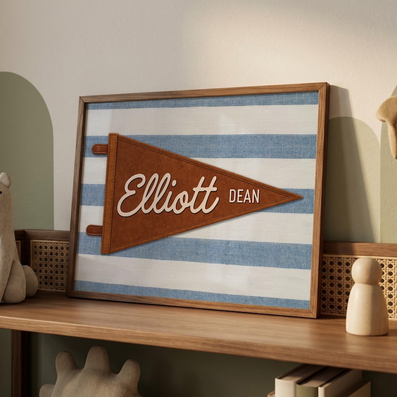 May include: A rectangular framed wall art piece featuring a brown felt pennant with the name Elliott Dean in white script and block lettering. The pennant is centered on a background of horizontal blue and white stripes within a wooden frame.