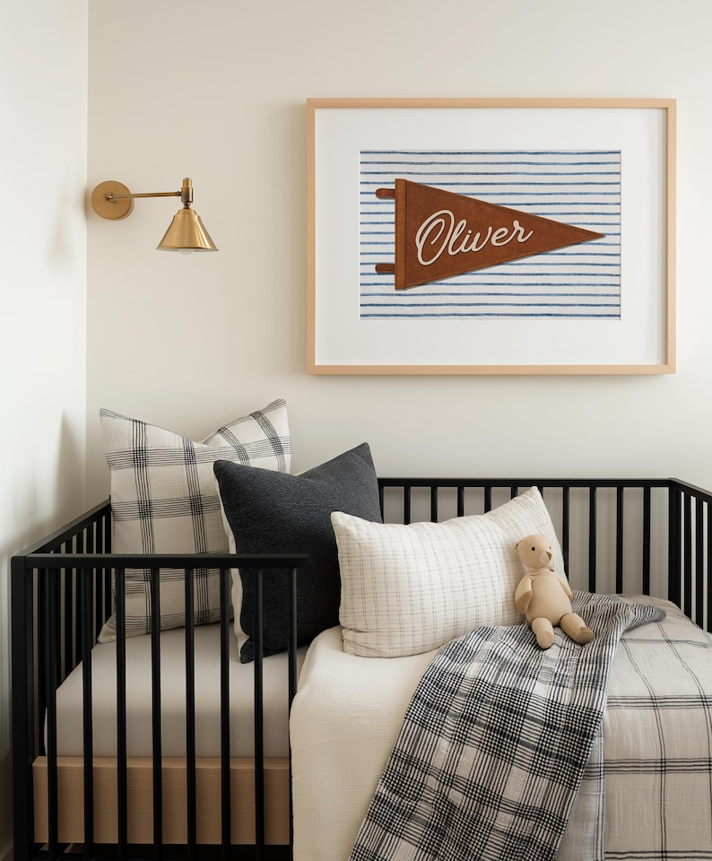 May include: A framed wall art print featuring a brown felt pennant with the name Oliver in white script, set against a blue and white striped background. The print is displayed in a light wood frame above a black crib with various pillows.