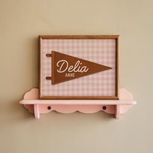 May include: A framed pennant with the name "Delia Anne" in a cursive font. The pennant is brown and set against a pink and white gingham background. The frame is wood, and the artwork is displayed on a pink, cloud-shaped shelf.