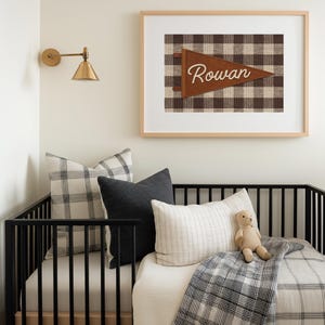 May include: A nursery scene featuring a black crib with pillows and a blanket in neutral tones. A framed print with the name "Rowan" on a pennant hangs above the crib. A gold sconce is mounted on the wall.