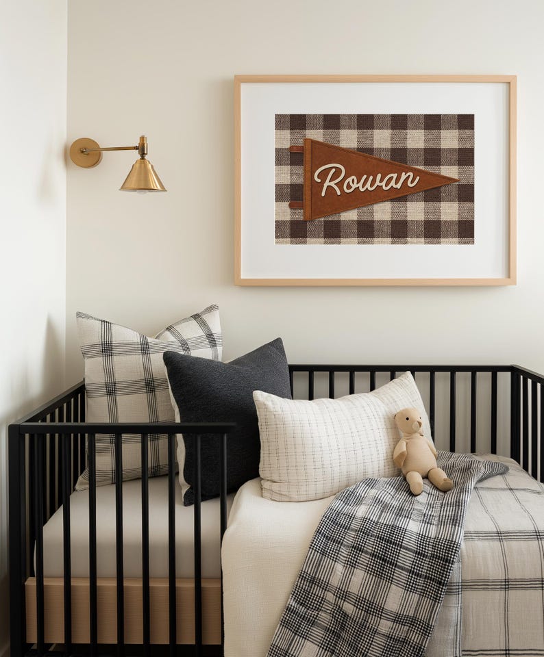 May include: A nursery scene featuring a black crib with pillows and a blanket in neutral tones. A framed print with the name "Rowan" on a pennant hangs above the crib. A gold sconce is mounted on the wall.