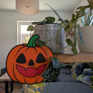 Könnte beinhalten: Eine orangefarbene Kürbisdekoration mit einem lächelnden Gesicht, schwarzen Augen und einem grünen Stiel. Die Halloween-Dekoration ist auf einer silbernen Stange befestigt. Im Hintergrund sind Topfpflanzen und eine Wohnzimmereinrichtung zu sehen.