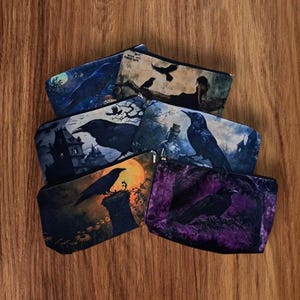 May include: A collection of zippered pouches with various raven-themed illustrations. The pouches feature images of ravens perched on tombstones, in front of moons, and near haunted houses. The color palette includes blues, purples, and oranges.