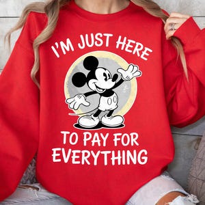 May include: Red sweatshirt featuring Mickey Mouse with the text "I'M JUST HERE TO PAY FOR EVERYTHING." Mickey Mouse is in black, white, and gray, set against a yellow and gray circle.