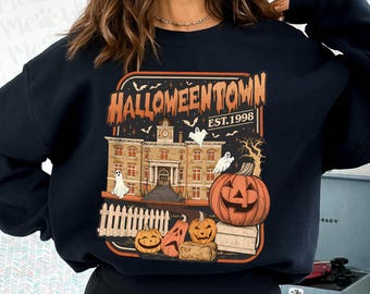 May include: Black sweatshirt featuring a Halloween-themed graphic. The design includes a building, pumpkins, ghosts, and the text "Halloweentown Est. 1998" in orange and white. A white picket fence is in the foreground.