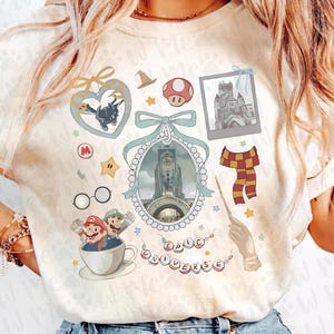 Puede incluir: Camiseta color crema con varios gráficos de dibujos animados. Presenta un diseño en forma de corazón con un dragón, una seta, una foto de un castillo, una bufanda y una varita. Las palabras "EPIC UNIVERSE" están escritas en letras individuales.