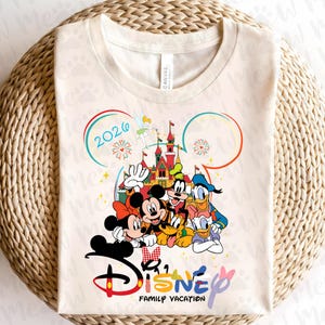 May include: Cream-colored t-shirt featuring a colorful Disney graphic. The design includes Mickey and Minnie Mouse, Donald Duck, Goofy, and Pluto in front of a castle. The text "Disney Family Vacation 2026" is also included.