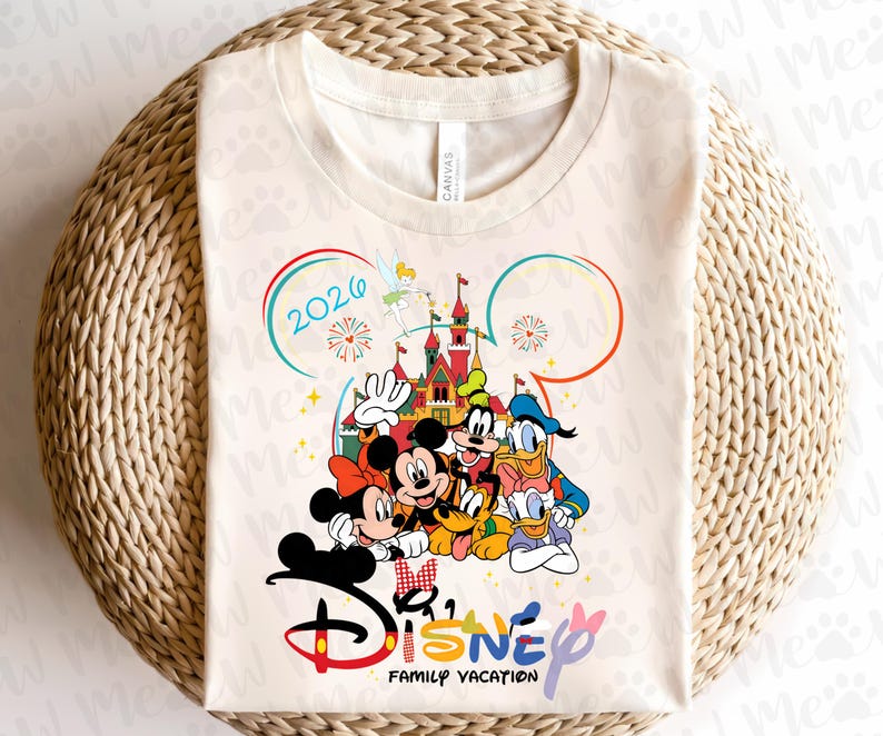 May include: Cream-colored t-shirt featuring a colorful Disney graphic. The design includes Mickey and Minnie Mouse, Donald Duck, Goofy, and Pluto in front of a castle. The text "Disney Family Vacation 2026" is also included.