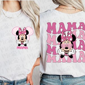 May include: White t-shirt featuring a pink Minnie Mouse graphic with a polka dot bow and the word "MAMA" in pink. The back of the shirt repeats "MAMA" in a retro font, with Minnie wearing heart-shaped sunglasses.