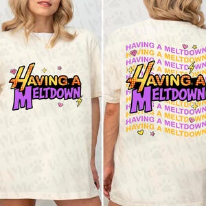 Puede incluir: Camiseta color crema con el texto "HAVING A MELTDOWN" en amarillo y morado, con un rayo y una estrella. La parte trasera de la camiseta repite la frase con gr&aacute;ficos adicionales.