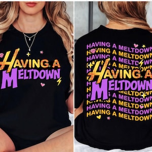 Puede incluir: Camiseta negra con la frase "HAVING A MELTDOWN" en amarillo, naranja y morado. El dise&ntilde;o incluye un diamante, un rayo y corazones. La parte trasera de la camiseta repite la frase.