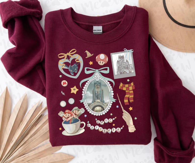 Pu&ograve; includere: Una felpa bordeaux con varie grafiche a tema cartoni animati. Il design include un cuore con un pipistrello, un fungo, un castello, una sciarpa, una bacchetta e personaggi di Mario in una tazza. &Egrave; presente anche il testo "Epic Universe".