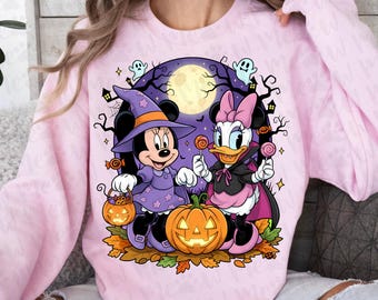 Könnte beinhalten: Ein hellrosa Sweatshirt mit einem Halloween-Motiv. Die Grafik zeigt Minnie Maus und Daisy Duck in Halloween-Kostümen, die Lutscher halten und sich in der Nähe eines geschnitzten Kürbisses aufhalten. Der Hintergrund umfasst einen Vollmond, Geister und gruselige Bäume.