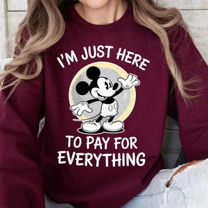May include: Burgundy sweatshirt featuring Mickey Mouse with the text "I'M JUST HERE TO PAY FOR EVERYTHING." Mickey Mouse is in black, white, and gray, standing in front of a yellow and gray circle.
