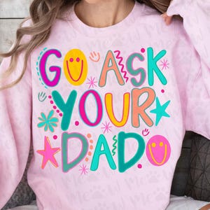 May include: A light pink sweatshirt with the words "GO ASK YOUR DAD" in colorful, playful lettering. The design includes smiley faces, stars, and floral accents, creating a fun and whimsical aesthetic. The sweatshirt is a comfortable, casual style.