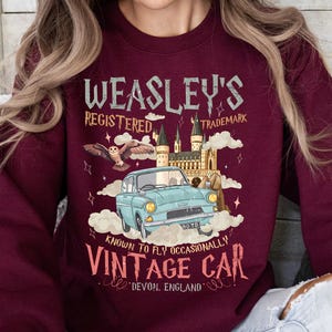 Puede incluir: Sudadera burdeos con un gr&aacute;fico de un coche antiguo, un b&uacute;ho y un castillo. El texto dice "Weasley's Registered Trademark" y "Vintage Car, Devon, England". El coche es de color azul claro.