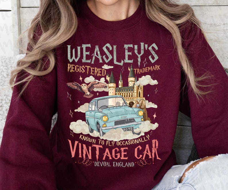 Puede incluir: Sudadera burdeos con un gr&aacute;fico de un coche antiguo, un b&uacute;ho y un castillo. El texto dice "Weasley's Registered Trademark" y "Vintage Car, Devon, England". El coche es de color azul claro.