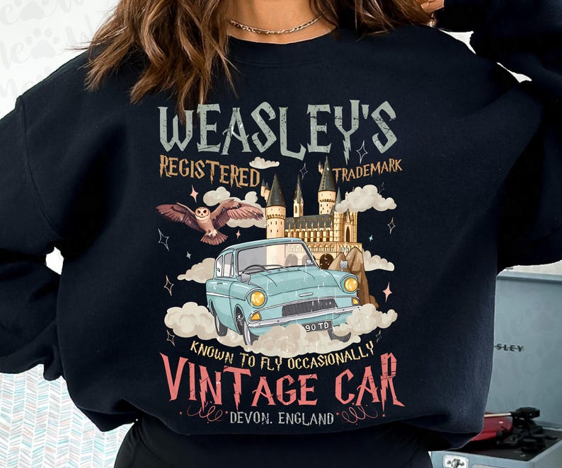 Puede incluir: Sudadera negra con un dise&ntilde;o de coche vintage. El texto dice "Weasley's Registered Trademark Vintage Car", con un b&uacute;ho, un castillo y la frase "Known to Fly Occasionally". El coche es de color azul claro.
