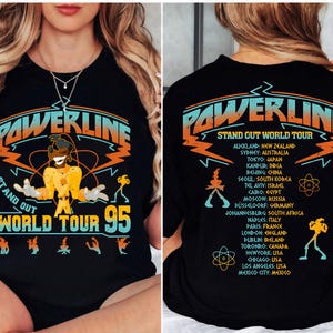 May include: Black t-shirt with the text "POWERLINE STAND OUT WORLD TOUR 95" in orange and teal. The front graphic includes a cartoon character. The back lists tour locations including Auckland, Sydney, Tokyo, and more.