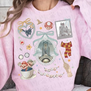 Pu&ograve; includere: Una felpa rosa chiaro con varie grafiche a cartoni animati. I disegni includono un cuore con un drago, un fungo, un castello, una sciarpa e la scritta "EPIC UNIVERSE". La felpa ha maniche lunghe e girocollo.