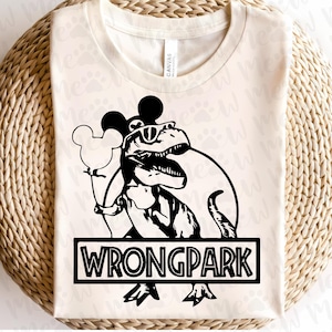 Puede incluir: Camiseta color crema con un gráfico negro de un dinosaurio con gafas de sol y orejas de Mickey Mouse, sosteniendo un globo de Mickey Mouse. El texto "WRONGPARK" se muestra en un cuadro rectangular negro debajo.