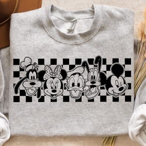 Op de afbeelding: Een lichtgrijs sweatshirt met een zwart-witte afbeelding van stripfiguren, waaronder Goofy, Minnie Mouse, Donald Duck, Pluto en Mickey Mouse. De figuren staan tegen een zwart-witte geblokte achtergrond.