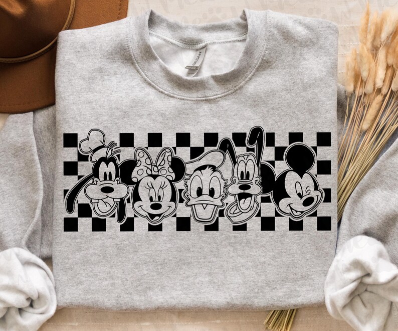 Op de afbeelding: Een lichtgrijs sweatshirt met een zwart-witte afbeelding van stripfiguren, waaronder Goofy, Minnie Mouse, Donald Duck, Pluto en Mickey Mouse. De figuren staan tegen een zwart-witte geblokte achtergrond.