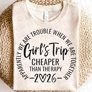 May include: Cream-colored t-shirt with the text "Girl's Trip CHEAPER THAN THERAPY -2026-" in black. The text is arranged in a circular design. The shirt is made of a soft material.
