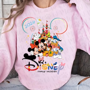 May include: A light pink sweatshirt featuring a colorful Disney design. The graphic includes Mickey and Minnie Mouse, Donald Duck, Goofy, and a castle. The text "Disney Family Vacation 2026" is also displayed.