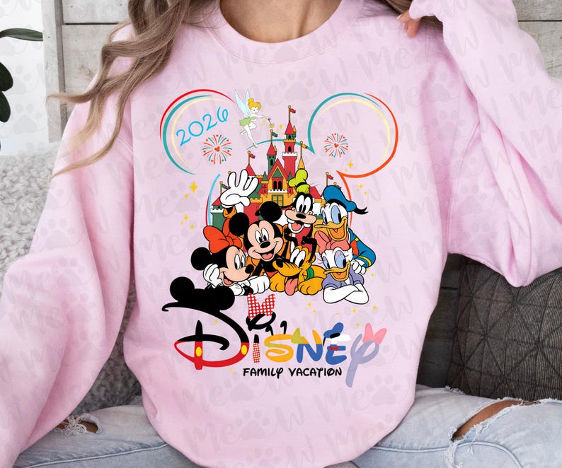 May include: A light pink sweatshirt featuring a colorful Disney design. The graphic includes Mickey and Minnie Mouse, Donald Duck, Goofy, and a castle. The text "Disney Family Vacation 2026" is also displayed.