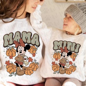 May include: White sweatshirts with matching "Mama" and "Mini" designs. Each design features Minnie Mouse in autumn attire, surrounded by pumpkins and leaves. The "Mama" design is larger than the "Mini" design.