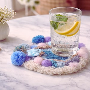 Puede incluir: Un vaso de agua clara con limón y menta descansa sobre un posavasos hecho a mano. El posavasos es redondo, con un diseño texturizado multicolor con pompones azules, morados y rosas. Un pequeño jarrón blanco con flores blancas está en el fondo.