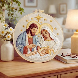 Puede incluir: Bordado circular de la Sagrada Familia: Jesús, María y el niño Jesús, con halos y estrellas. El bordado está en un aro de madera, con puntadas detalladas sobre un fondo crema. La obra de arte se exhibe sobre una superficie de madera.