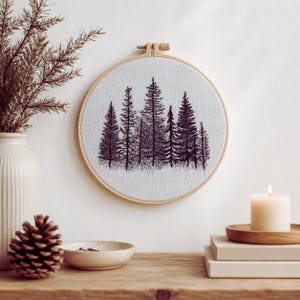 Può includere: Un telaio da ricamo circolare in legno contenente un tessuto grigio chiaro con un design dettagliato cucito a filo nero di una fitta foresta con molteplici alberi sempreverdi di varie altezze allineati al centro.