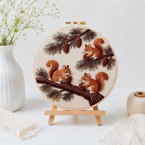 Può includere: Ricamo circolare con tre scoiattoli marroni su un ramo con pigne e aghi di pino. L'opera d'arte è incorniciata in un cerchio di legno ed è esposta su un piccolo cavalletto di legno. Lo sfondo è color crema.