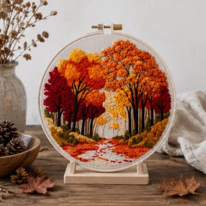 Könnte beinhalten: Ein rundes Stickbild im Stickrahmen zeigt eine herbstliche Waldszene mit texturierten Fäden in leuchtendem Rot, Orange und Gelb, die Bäume und einen gewundenen Pfad darstellen. Die Stickerei steht auf einem hellen Holzständer.