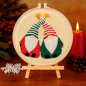 Op de afbeelding: Een geborduurde afbeelding met twee kabouters in rode en groene outfits en gestreepte hoeden. De kabouters hebben witte baarden en gele pompons op hun hoeden. De borduurwerk wordt weergegeven op een houten ezel, met een kaars en een dennenappel op de achtergrond.