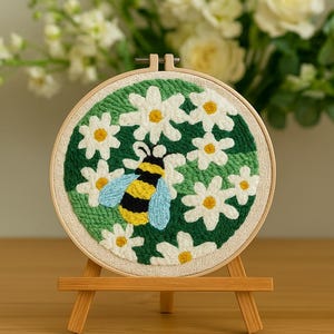 Op de afbeelding: Geborduurde hoepelkunst met een bij en madeliefjes. De bij heeft gele en zwarte strepen met blauwe vleugels, omringd door witte madeliefjes met gele centra op een groene achtergrond. De borduursels worden gepresenteerd op een klein houten ezel.
