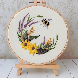 Könnte beinhalten: Gestickte Blumenkunst in einem Stickrahmen mit einer Biene, gelben Sonnenblumen und lila Lavendel. Das Design befindet sich auf einem cremefarbenen Stoff in einem Holzrahmen, der auf einer kleinen Holzstaffelei ausgestellt ist. Die Stickerei ist detailliert und farbenfroh.