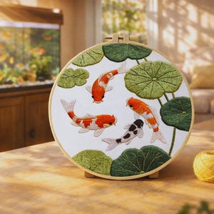 May include: Round embroidery hoop featuring a detailed design of koi fish and lily pads. The fish are in shades of orange, white, and black, with green lily pads. The embroidery is set against a white background.
