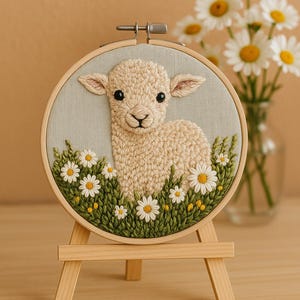 Op de afbeelding: Handgeborduurde kunst met een lammetje in een veld met madeliefjes. Het lammetje is beige met zwarte ogen, omringd door groen gras en witte madeliefjes met gele centra. Het kunstwerk wordt gepresenteerd in een houten borduurring op een houten ezel.