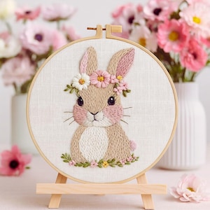 May include: Embroidered bunny hoop art. The artwork features a tan bunny with pink accents, wearing a floral crown. The bunny is surrounded by a floral border and set in a wooden hoop. The hoop is displayed on a small wooden easel.