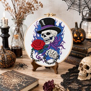 May include: A circular embroidery hoop displaying a stitched design of a skull wearing a black top hat with a purple band. The skull holds a red rose and is surrounded by blue flames and a grey chain, all set against a white fabric background.