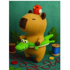 Puede incluir: Un juguete de capibara tejido con un cocodrilo verde envuelto alrededor de su cuerpo. Un pequeño pájaro rojo está sentado en la cabeza del capibara. La palabra "Capibara" está escrita en verde encima del juguete. Suministros de costura, incluyendo hilo, botones y una aguja, están en el fondo.